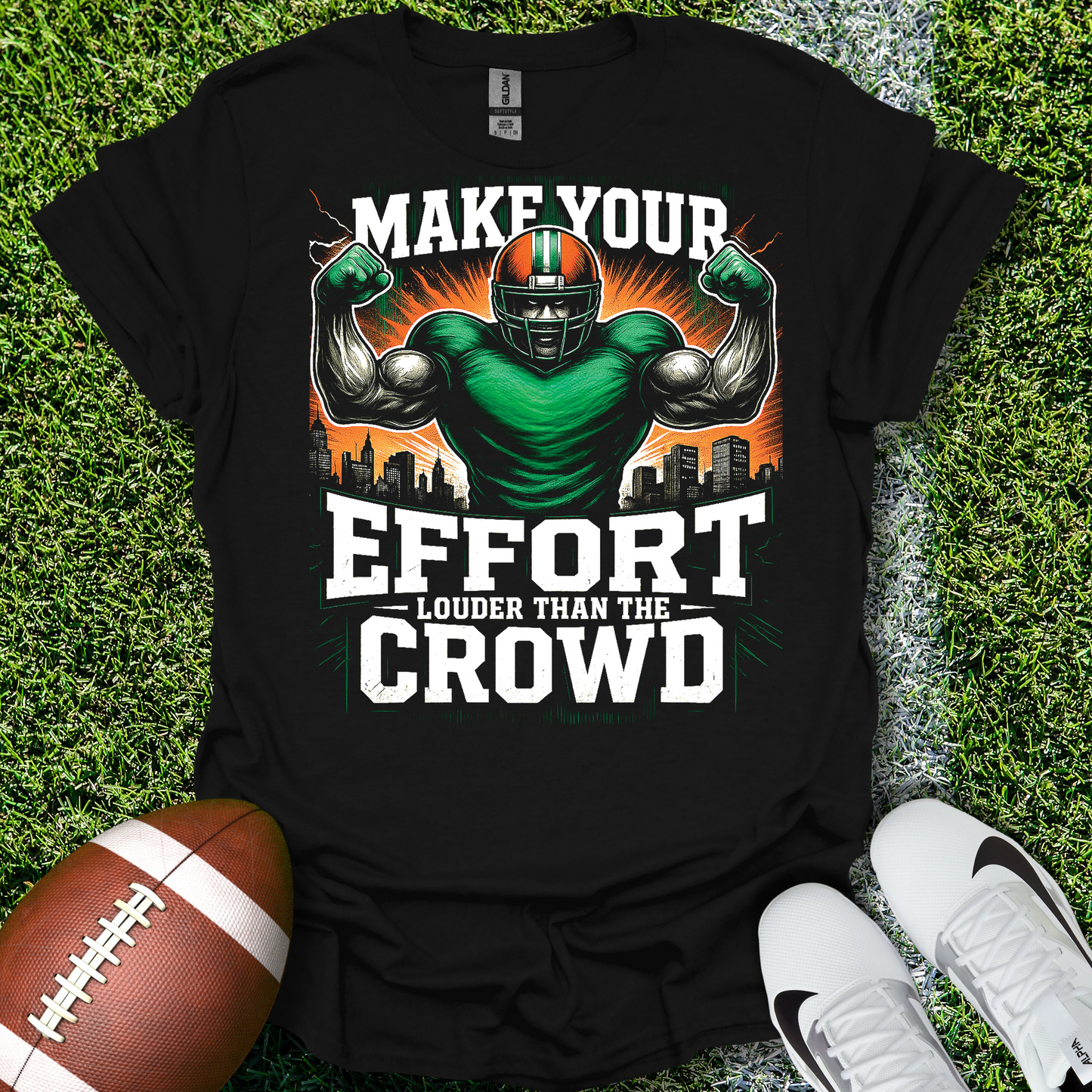 Black t-shirt with sports-themed graphic and text on grass with football and shoes.