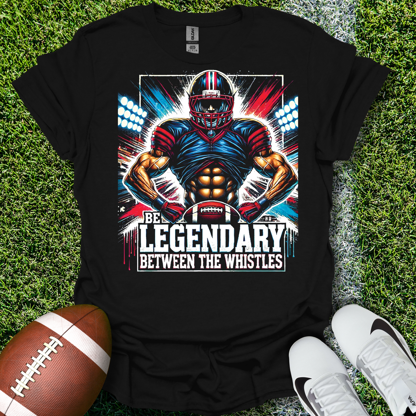Black t-shirt with football player graphic and text 'Be Legendary Between the Whistles' on grass with a football and shoes.