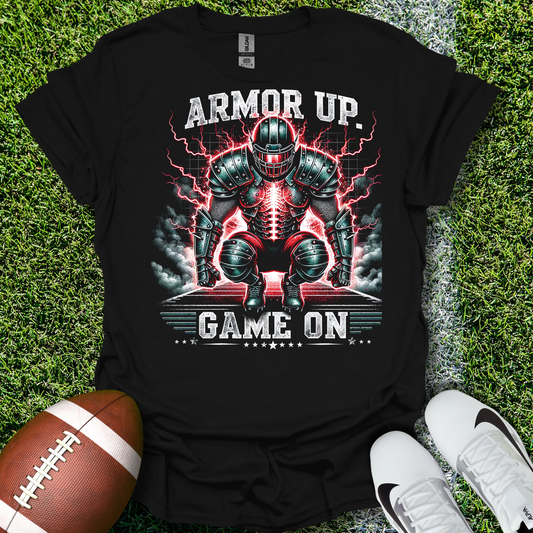 Black t-shirt with a football player in armor graphic and 'Armor Up Game On' text on grass with a football and shoes.