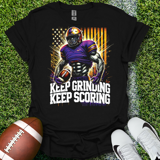 Black t-shirt with football player graphic and 'Keep Grinding Keep Scoring' text on grass with a football and shoes.