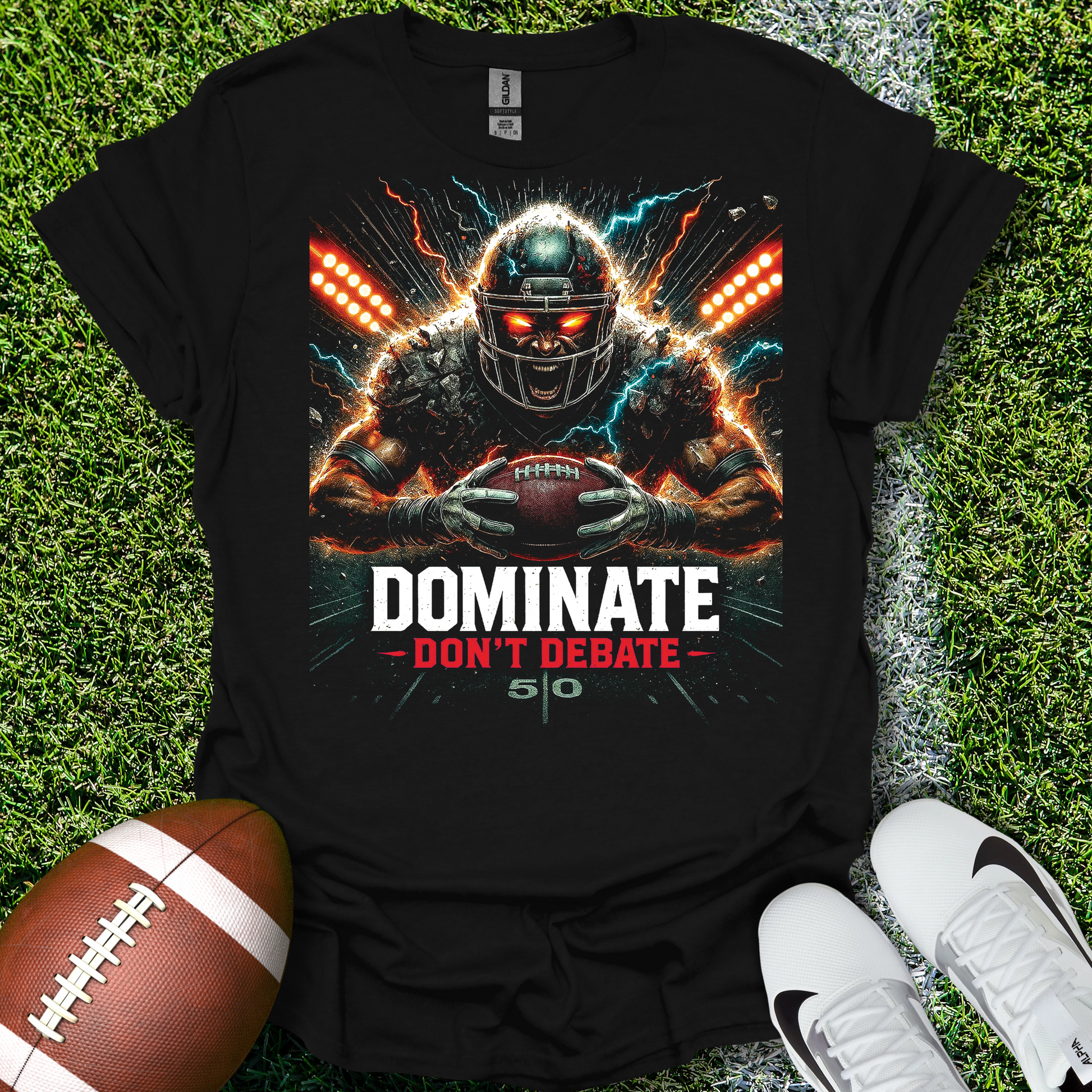 Black t-shirt with football player graphic and text 'DOMINATE DON'T DEBATE' on grass with a football and shoes.